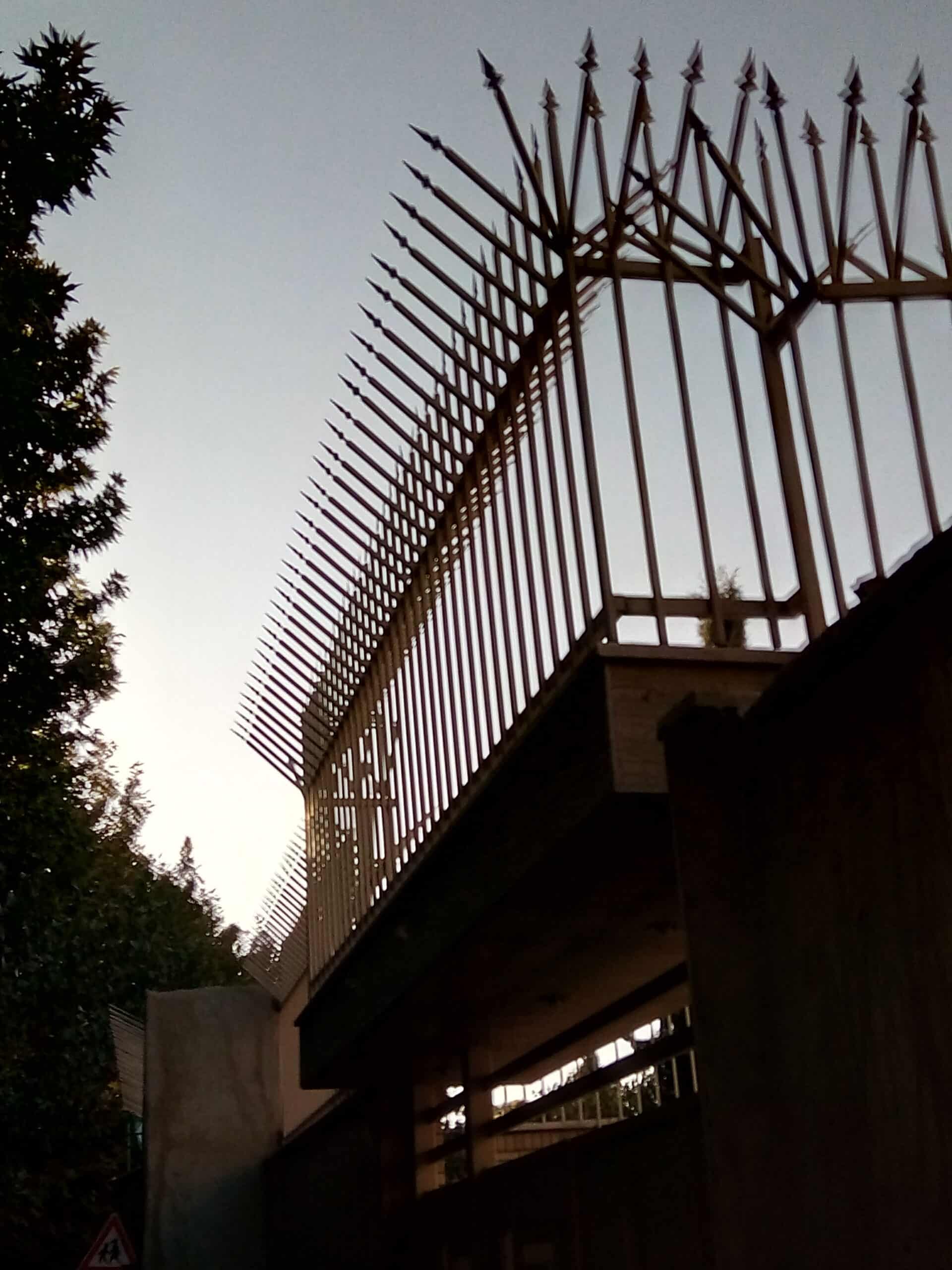 Fence_in_Iran_to_protect_a_house_(2017) Cerca de metal com hastes em 45º no topo, num céu cinzento