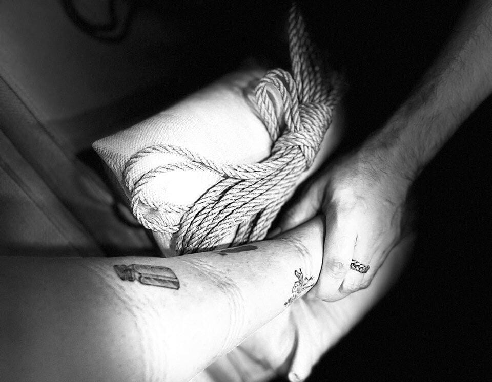 trama imagem de uma mão segurando uma perna com tatuagens, com um rolo de cordas ao lado, apoiados em uma almofada.